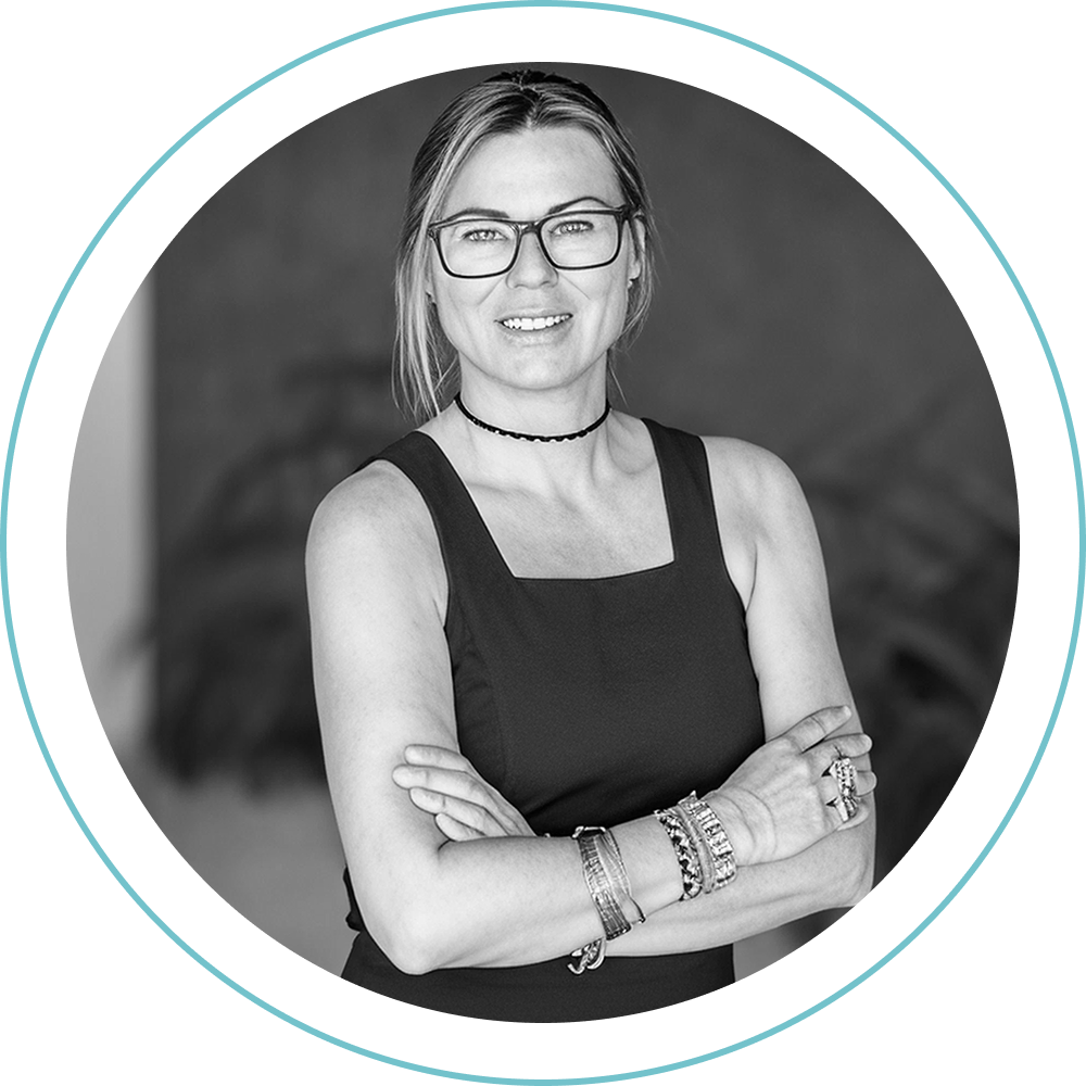 Una mujer con gafas y el pelo largo recogido hacia atrás sonríe con los brazos cruzados. Lleva un top sin mangas, un collar gargantilla y varias pulseras. El retrato es en blanco y negro con un borde circular.