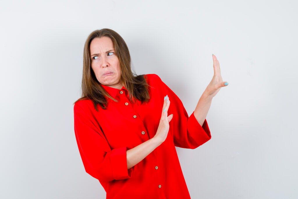 Una mujer con camisa roja, de pie sobre un fondo blanco liso, mira a su izquierda con expresión de disgusto y levanta las manos como para protegerse, tal vez reaccionando a las preocupaciones de El nuevo consumidor energético.