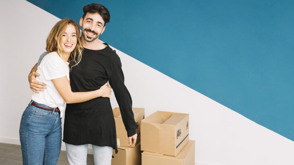 Una pareja sonriente se abraza frente a una pared azul y blanca, con varias cajas de cartón de mudanza apiladas cerca, encarnando al nuevo consumidor energético mientras se mudan a una nueva casa energéticamente eficiente.