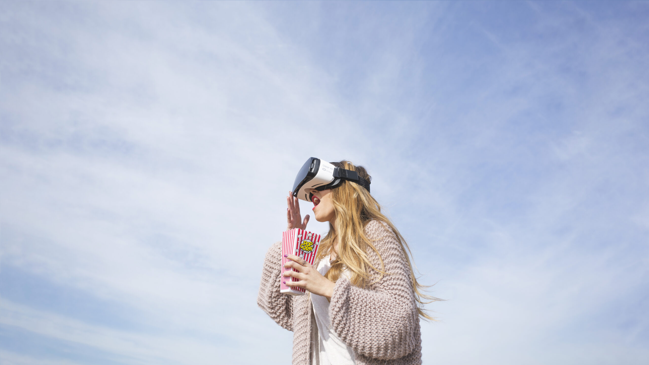 Una mujer con un casco de realidad virtual sostiene una caja de palomitas a rayas, al aire libre, bajo un cielo azul con nubes difusas, en la Loyalty Experience - Madrid Summit 2025.
