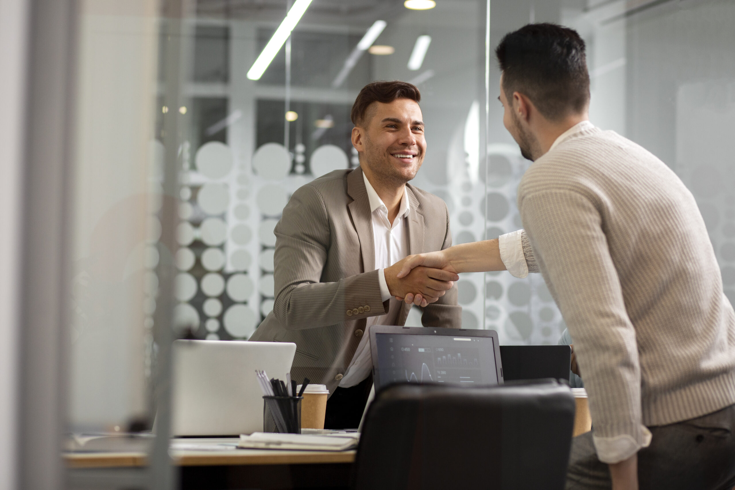 Dos hombres se dan la mano sonrientes en un despacho. Sobre la mesa, colocada contra una pared de cristal con motivos circulares, se ven ordenadores portátiles, documentos y material de oficina, que captan el espíritu de colaboración de la Loyalty Experience - Madrid Summit 2025.