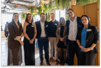 Un grupo de siete personas vestidas profesionalmente permanecen de pie en el interior, sonriendo a la cámara. El escenario con suelos de madera, luz natural y plantas verdes colgantes invita a conocer nuestro Loyalty Hub.