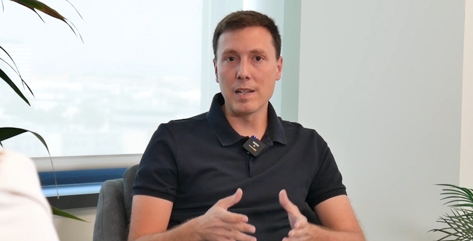 Una persona de pelo castaño corto, con un polo negro y un micrófono, está sentada en el interior y habla, gesticulando con ambas manos. Detrás de ellos hay plantas verdes y una ventana, perfecta para las ponencias de nuestros expertos para profesionales que quieren ir un paso más allá.