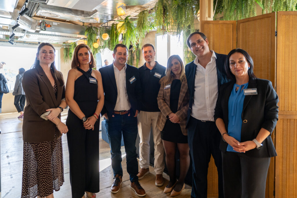 Siete personas en traje de negocios permanecen juntas en el interior, sonriendo a la cámara. El escenario presenta suelos de madera, vegetación colgando del techo y luz natural que entra por la izquierda. Conoce nuestro Loyalty Hub en un ambiente profesional y acogedor.