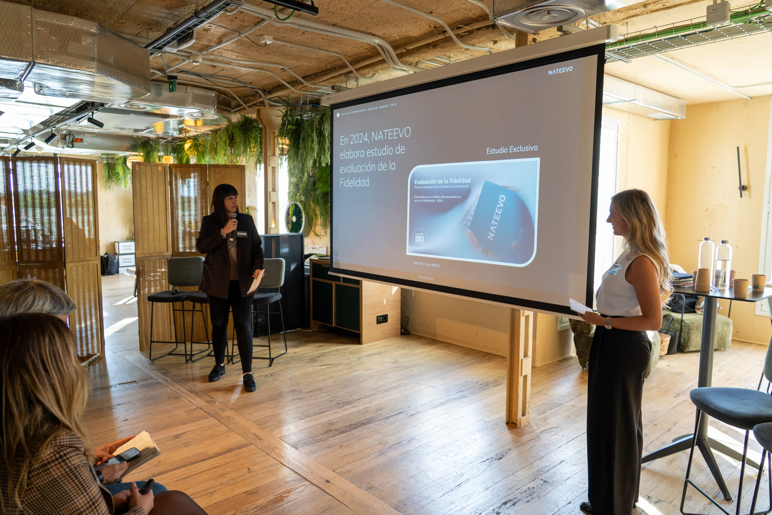 Dos mujeres se sitúan al frente de una sala moderna y bien iluminada, presentando información en la pantalla de un proyector sobre Loyalty Hub: Innovación e hiperpersonalización en la era digital y un estudio NATEVO 2024 a un público sentado.