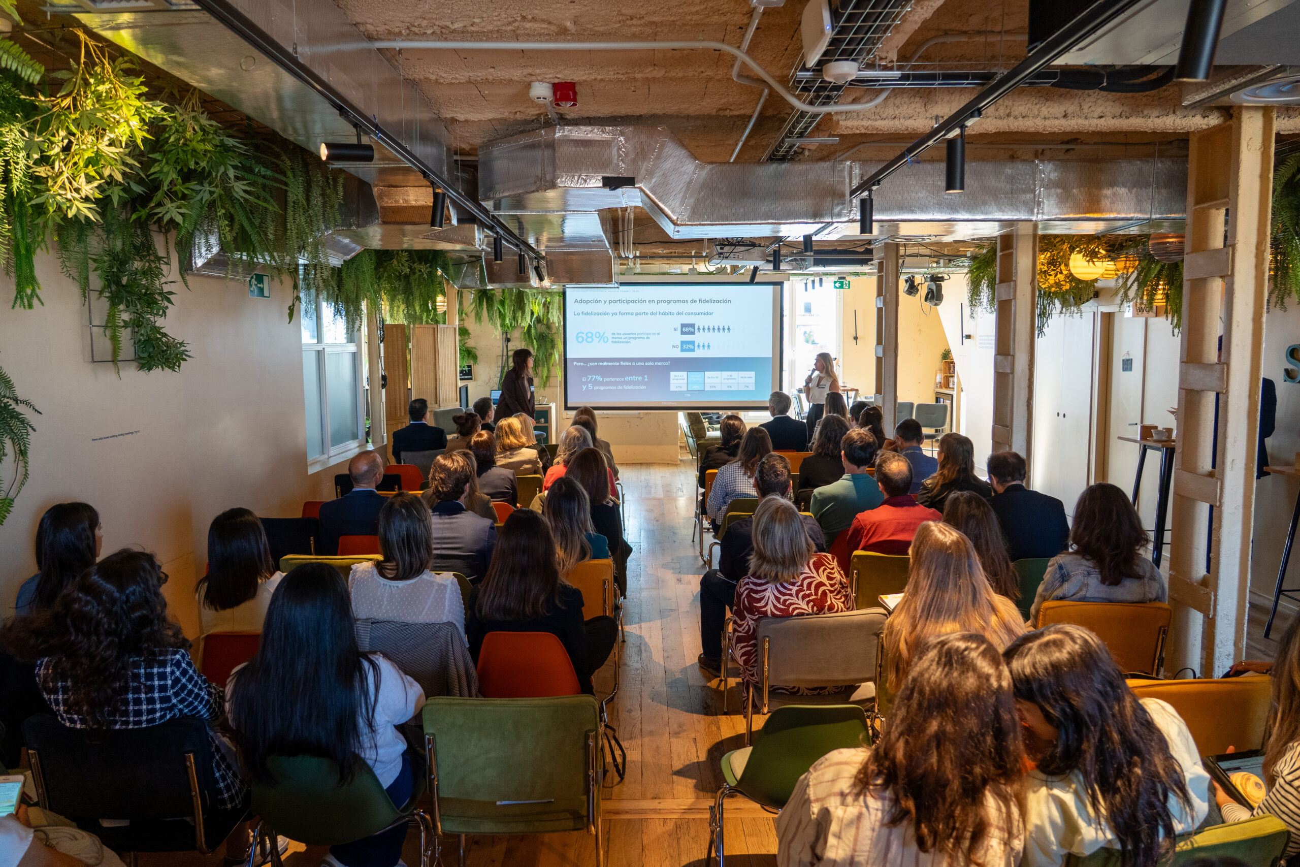 Un grupo de personas sentadas frente a una gran pantalla en una sala moderna y llena de plantas asisten a un seminario sobre Loyalty Hub: Innovación e hiperpersonalización en la era digital. La pantalla muestra diapositivas con gráficos y texto.