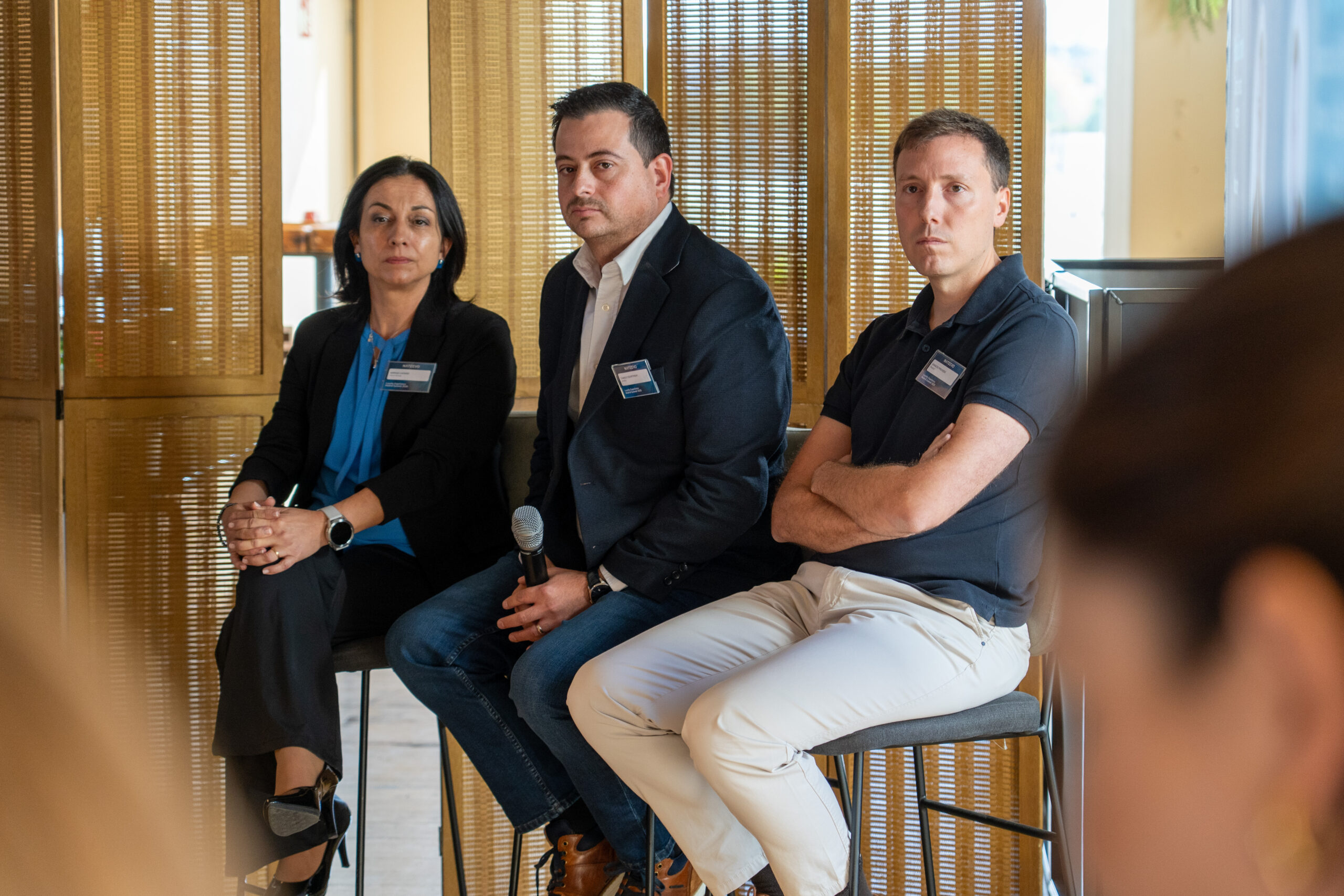 Tres personas sentadas en taburetes en una mesa redonda, vestidas con ropa informal de negocios y etiquetas con su nombre. Una de ellas sostiene un micrófono y mantiene una conversación sobre Loyalty Hub: Innovación e hiperpersonalización en la era digital, con pantallas de madera de fondo.