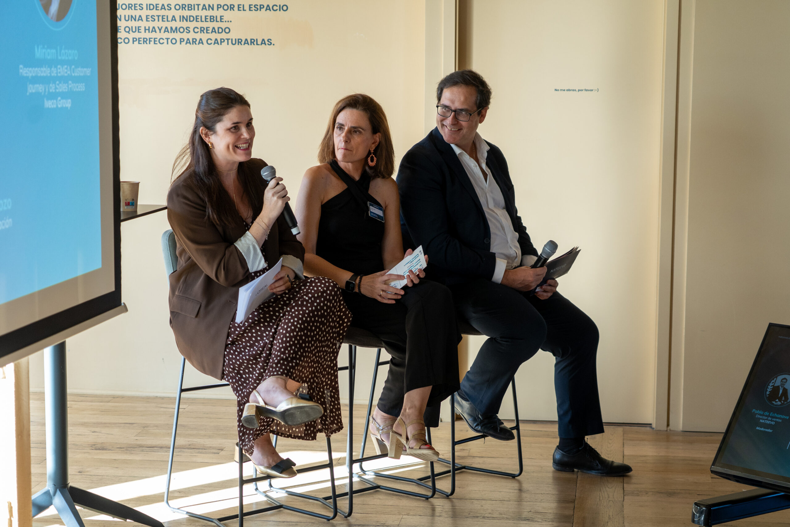Tres personas sentadas en sillas altas en el interior durante una mesa redonda sobre Loyalty Hub: Innovación e hiperpersonalización en la era digital. La mujer de la izquierda habla por un micrófono, mientras los demás escuchan y sostienen papeles. Se ve una pantalla de presentación.