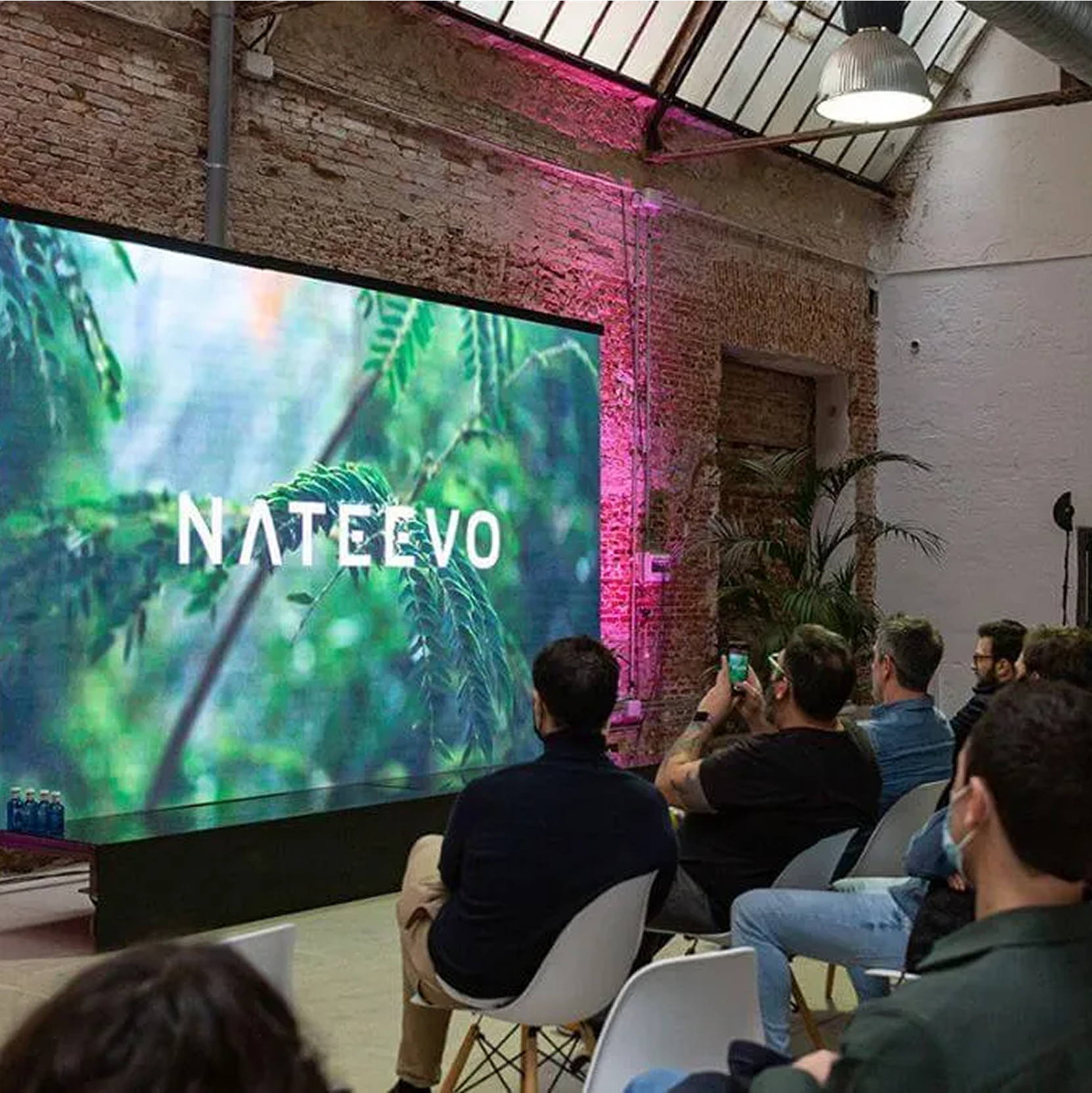 Los asistentes se sientan en sillas frente a una gran pantalla en la que aparece la palabra NATEEVO, rodeados de vegetación, en el interior de un local tipo loft con paredes de ladrillo visto: un entorno inspirador que pone de relieve el cuidado diseño de la experiencia.