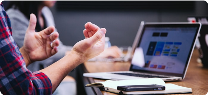 Una persona gesticula sentada a una mesa con un ordenador portátil, un cuaderno y un teléfono, inmersa en el diseño de experiencias. Una figura borrosa al fondo sugiere una reunión o un entorno de trabajo colaborativo.