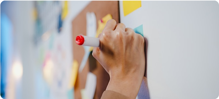 La mano de una persona sujeta un rotulador de pizarra blanca con punta roja, dedicada al diseño de experiencias, escribiendo o dibujando en una pizarra blanca con notas adhesivas de colores y papeles de fondo.