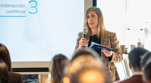 Una mujer con un micrófono y notas se dirige al público durante una presentación sobre simplicidad en el diseño UX. Una pantalla de proyección detrás de ella muestra el texto