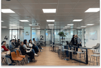 Un grupo de personas se sienta frente a dos presentadores en la parte delantera de una moderna sala de reuniones de oficina, donde se está debatiendo sobre IA como copiloto en marketing. Unas paredes de cristal numeradas y un proyector en la parte superior crean un ambiente profesional y atento.