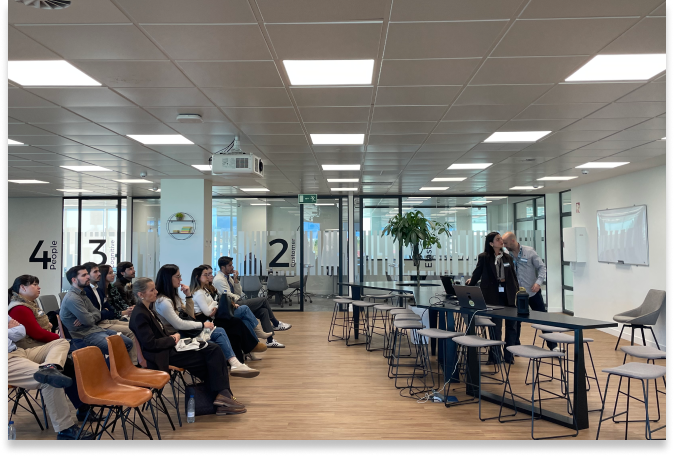 Un grupo de personas se sienta frente a dos presentadores en la parte delantera de una moderna sala de reuniones de oficina, donde se está debatiendo sobre IA como copiloto en marketing. Unas paredes de cristal numeradas y un proyector en la parte superior crean un ambiente profesional y atento.