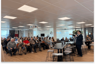 Un hombre trajeado habla y gesticula durante una presentación sobre Acciones Brand2Brand de manera ágil ante un público sentado en una sala de conferencias moderna y bien iluminada con grandes ventanales y un proyector montado en el techo.