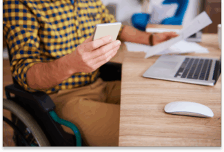 Una persona en silla de ruedas se sienta en un escritorio utilizando un smartphone y sosteniendo papeles, promoviendo la accesibilidad en proyectos digitales, con un ordenador portátil y un ratón de ordenador sobre el escritorio. Solo se ven el torso y las manos de la persona.