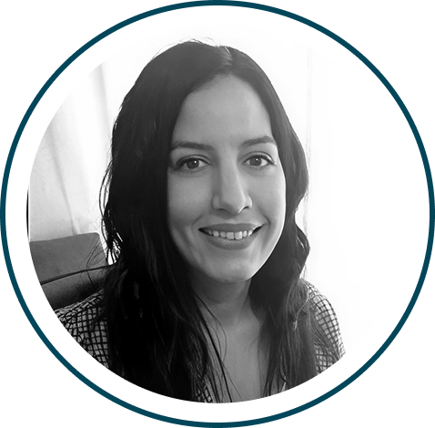 Fotografía en blanco y negro de una mujer con pelo largo y oscuro, vestida con un top a cuadros, que sonríe a la cámara. Está sentada con un fondo claro y la imagen está enmarcada por un borde circular.