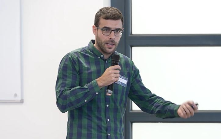 Un hombre con gafas y camisa de cuadros verde comparte casos de éxito de fidelización mientras habla por un micrófono y gesticula con la otra mano en una sala luminosa con un gran ventanal a sus espaldas.
