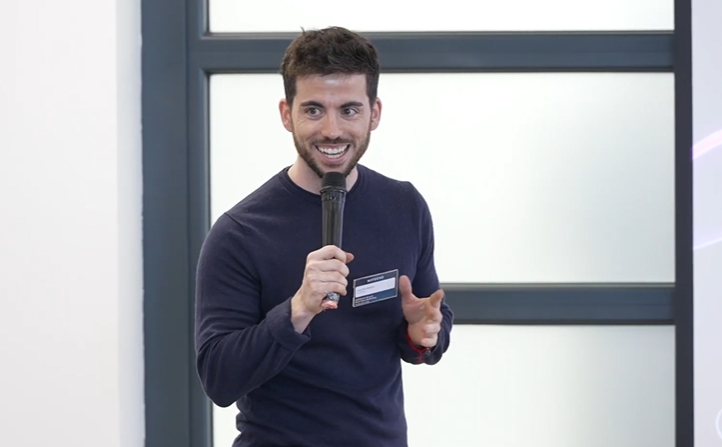 Un hombre de pelo corto y oscuro, con jersey azul marino y etiqueta con su nombre, sonríe y habla por un micrófono sobre Casos de éxito fidelización mientras gesticula con la mano. Está de pie frente a una ventana con paneles de cristal esmerilado.