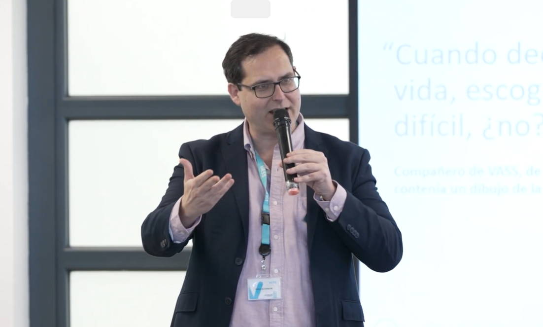 Un hombre con gafas y traje de chaqueta habla por un micrófono y gesticula con la mano. Se encuentra ante un gran ventanal y una pantalla que muestra un texto en español sobre Casos de éxito de fidelización.