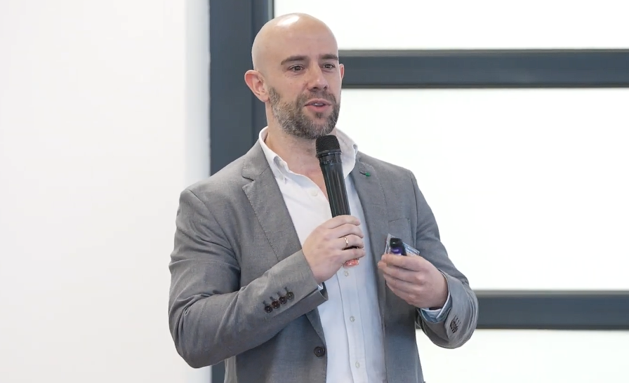 Un hombre calvo con barba, vestido con traje gris y camisa blanca, habla por un micrófono mientras sujeta un pequeño dispositivo durante su presentación en el Brunch de NATEEVO sobre marketing en tiempo real.