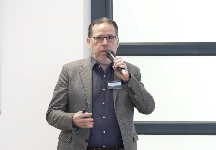 Un hombre con gafas y americana gris habla por un micrófono de mano mientras permanece de pie en el interior, gesticulando con la otra mano mientras comparte Casos de éxito fidelización con el público. Lleva el pelo corto y lleva barba, y está de cara al público.