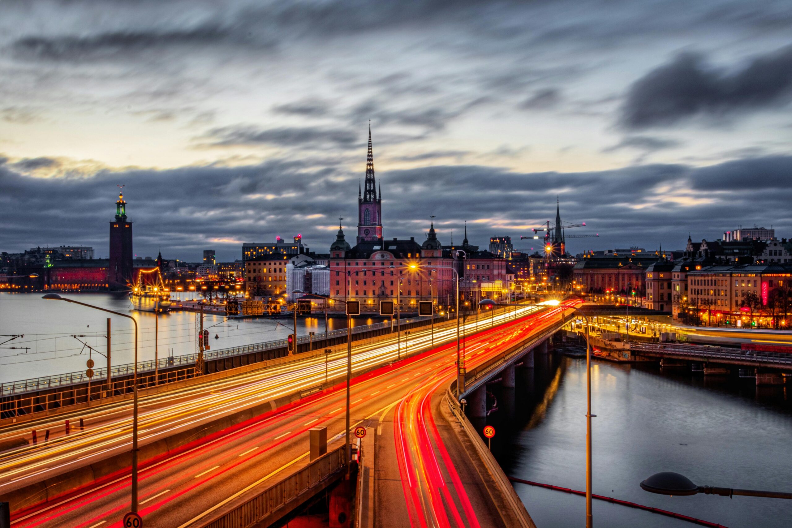 Foto de larga exposición de un paisaje urbano al atardecer, con estelas de luz brillante de vehículos en un puente, edificios históricos, una alta aguja de iglesia y agua que refleja las luces de la ciudad bajo un cielo nublado.
