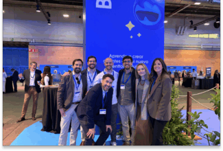 Siete personas posan y sonríen juntas delante de un gran cartel azul en un acto en interior centrado en los codazos y la economía conductual. Otros asistentes se mezclan en el fondo, y las mesas y la decoración del evento contribuyen a crear un ambiente animado.