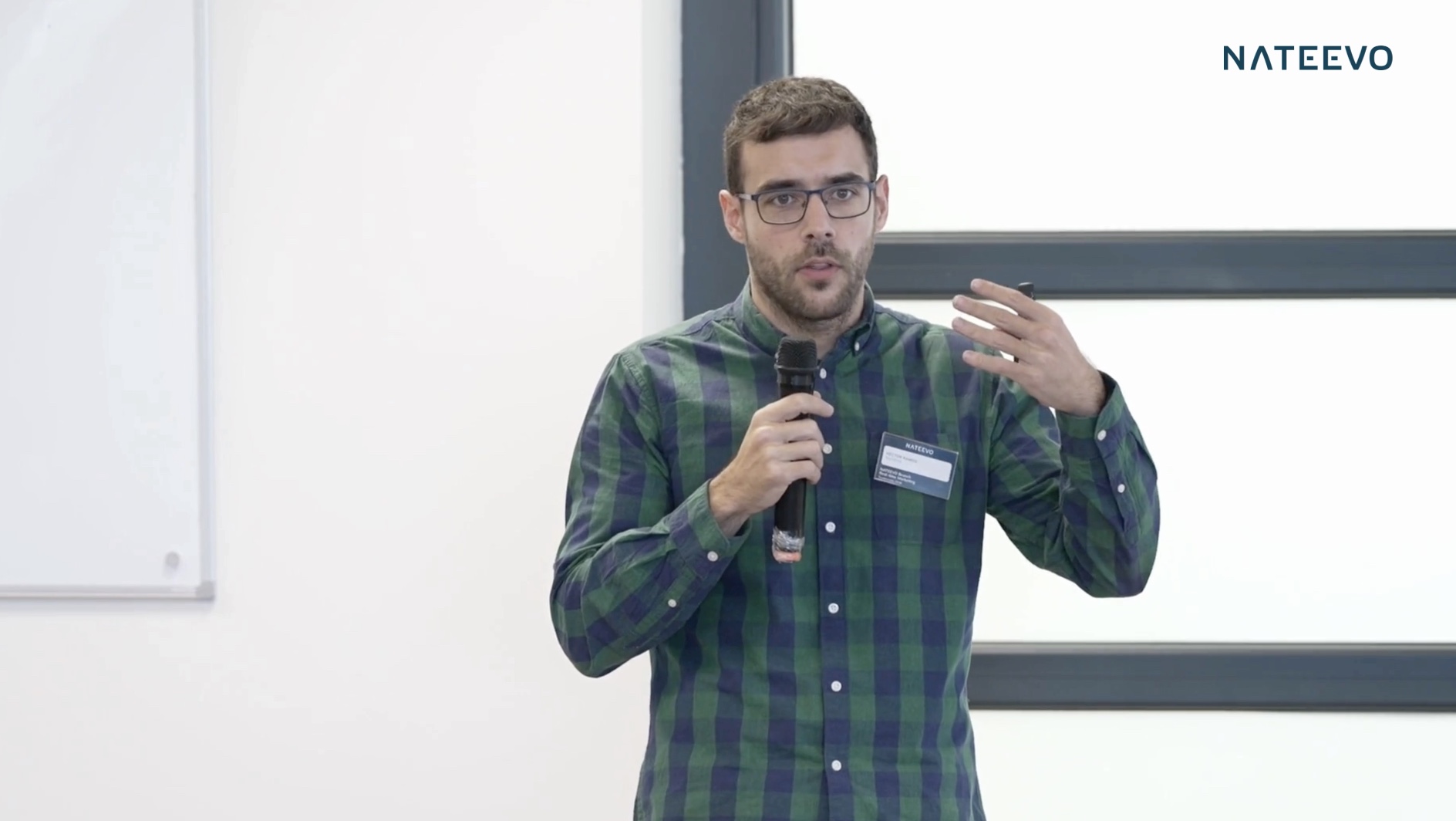 Un hombre con gafas y camisa verde a cuadros sostiene un micrófono y gesticula mientras habla delante de un fondo blanco con el logotipo NATEEVO en la esquina superior derecha.