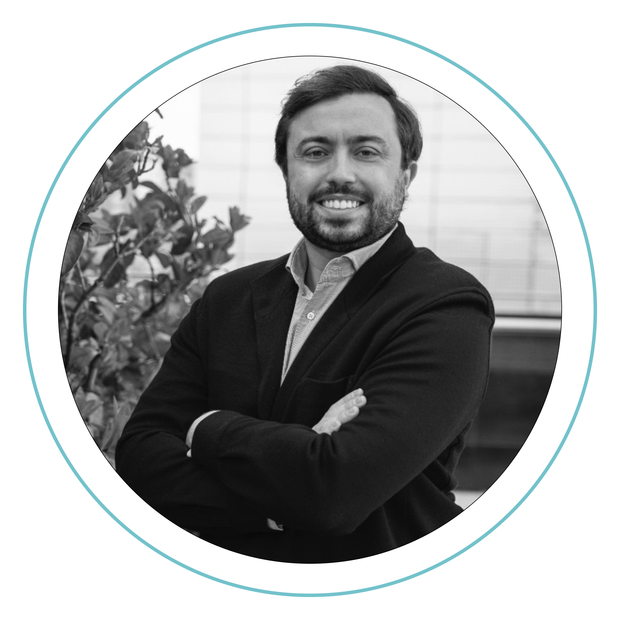 Fotografía en blanco y negro de un hombre con pelo corto y barba, sonriendo con los brazos cruzados, vestido con americana y camisa. Está de pie cerca de una planta, con un edificio moderno de fondo. Borde circular alrededor de la imagen.