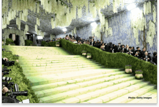 Una amplia y gran escalinata bordeada de frondosos setos verdes y plantas en macetas está rodeada de fotógrafos en la inteligencia artificial de la Met Gala 2026. Flores blancas cuelgan del techo, creando un ambiente elegante y ajardinado.