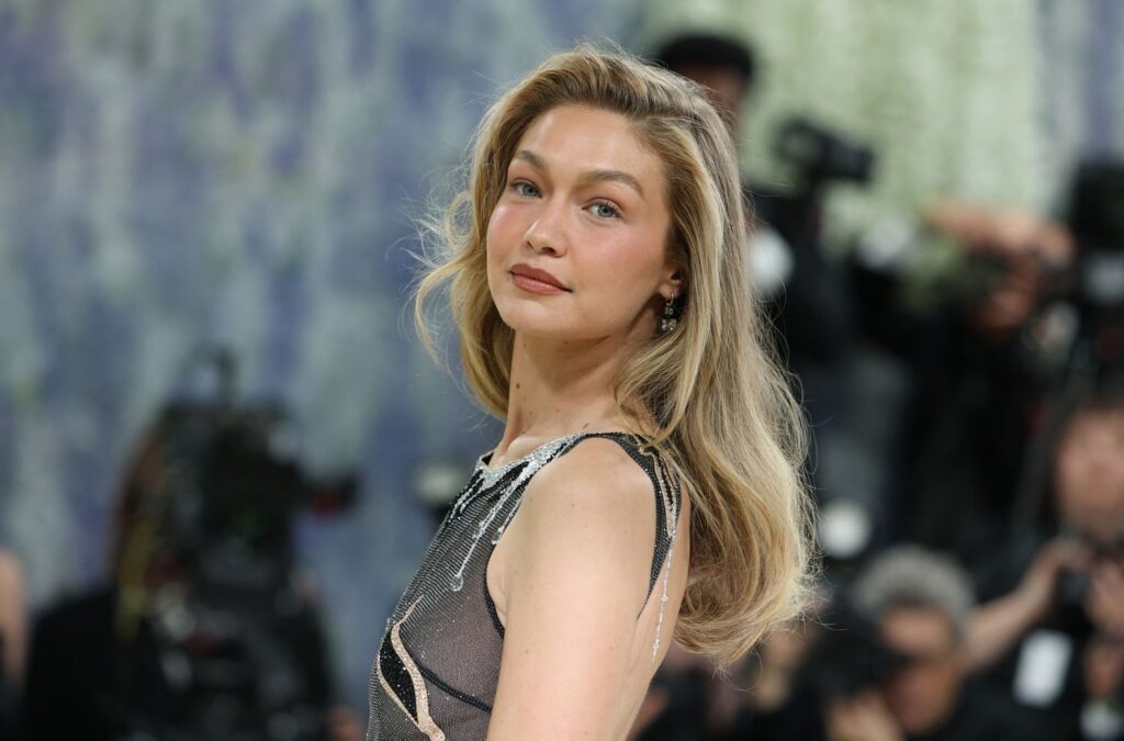 Una mujer con el pelo largo y rubio en ondas sueltas posa con un vestido sin mangas en la alfombra roja de la Met Gala 2026, mientras los fotógrafos captan su mirada y el zumbido de inteligencia artificial llena el fondo borroso.