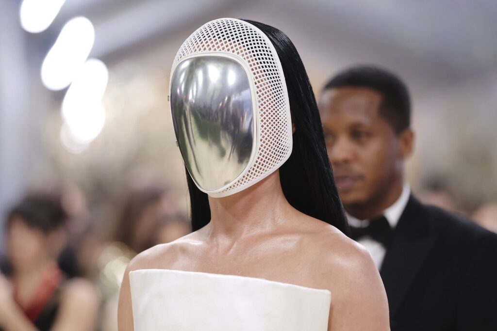 Una persona con una futurista máscara reflectante plateada con un marco de malla blanca y un traje blanco sin hombros se encuentra en un ambiente formal en la Gala Met 2026, donde los toques de inteligencia artificial inspiran a la multitud difuminada en el fondo.