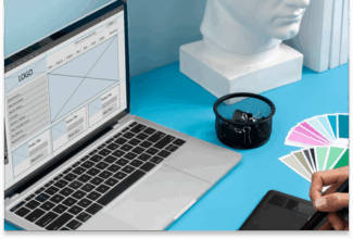 Una persona utiliza una tableta digital junto a un ordenador portátil en el que se muestra el diseño de una página web, con muestras de colores, un portalápices y una cabeza esculpida en blanco sobre un escritorio azul: un espacio de trabajo inspirador donde el diseño especulativo con IA da lugar a ideas innovadoras.