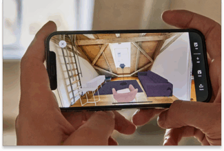 Una persona sostiene un smartphone en el que se ve una aplicación de diseño de interiores que muestra la personalización a escala, con una habitación virtual en la que destacan unas vigas de madera, un sofá morado, una silla rosa, un escritorio y una ventana al fondo.