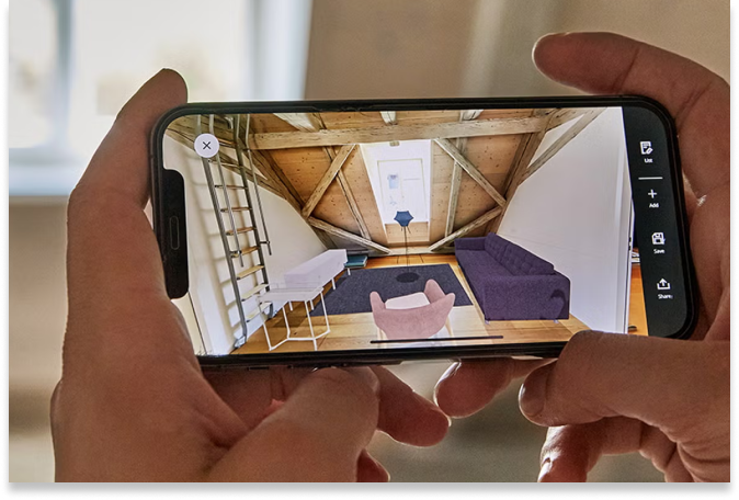 Una persona sostiene un smartphone en el que se ve una aplicación de diseño de interiores que muestra la personalización a escala, con una habitación virtual en la que destacan unas vigas de madera, un sofá morado, una silla rosa, un escritorio y una ventana al fondo.