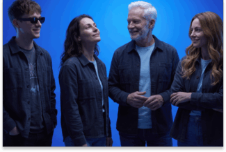 Cuatro personas posan juntas sobre un fondo azul, sonriendo y charlando. Tres mujeres y un hombre mayor, todos con chaquetas vaqueras y ropa informal, se muestran alegres y relajados, lo que refleja el ambiente amistoso y de conexión que se espera entre los consumidores de 2028.