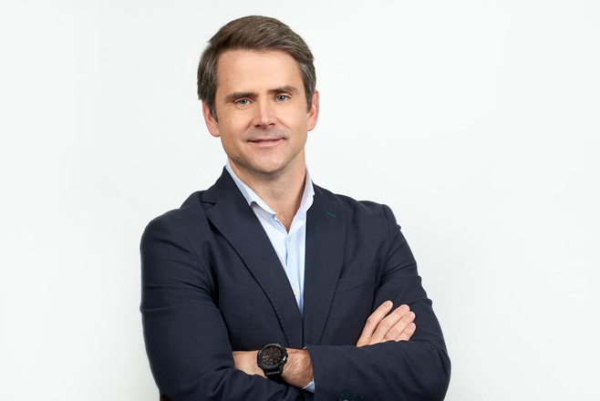 Un hombre con el pelo castaño corto, que lleva una chaqueta azul marino, una camisa azul claro y un reloj de pulsera, aparece de pie con los brazos cruzados y sonriendo a la cámara sobre un fondo blanco liso, encarnando la confianza y el espíritu de la personalización a escala.