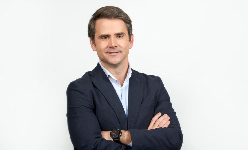 Un hombre con chaqueta de traje oscura y camisa azul claro aparece de pie, con los brazos cruzados y sonriendo, frente a un fondo blanco liso, encarnando la confianza y el profesionalismo —ideales para destacar la importancia de la personalización a gran escala en cualquier entorno empresarial—.