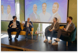 Cuatro hombres están sentados en un escenario durante una mesa redonda sobre la fidelización de clientes, frente a una pantalla en la que se muestran los retratos de los ponentes.